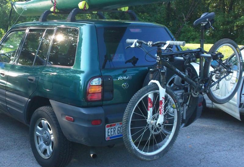 Gary Fisher Wahoo: A green Toyota RAV4 parked with a kayak secured on the roof rack and two bicycles mounted on the rear bike rack. The scene is set against a backdrop of greenery, suggesting an outdoor recreational environment.