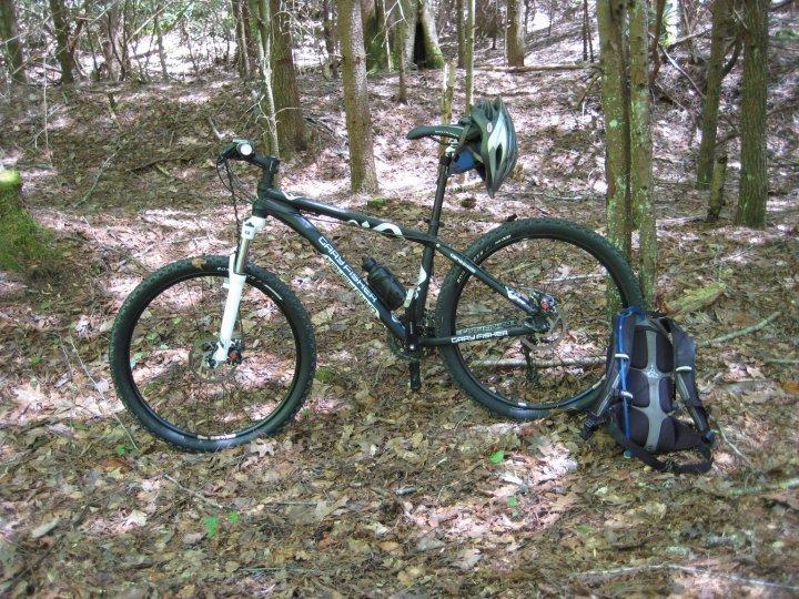 Gary Fisher Wahoo: A mountain bike with a helmet resting on the seat and a backpack on the ground beside it, located in a wooded area with trees and fallen leaves.