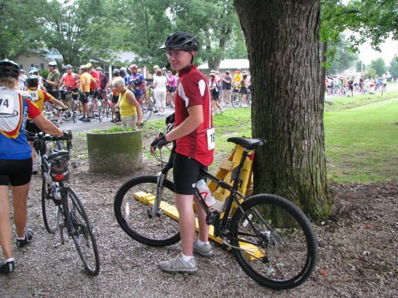 Gary Fisher Wahoo: A young cyclist wearing a red and white jersey and helmet stands beside a bicycle, smiling at the camera. In the background, a group of cyclists and spectators are gathered in a park setting, with trees and a pathway visible.