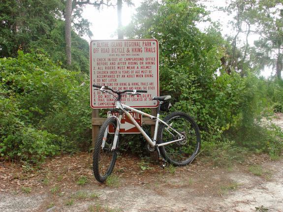 Gary Fisher X-Caliber: Mountain bike leaning against a sign for Blount Island Regional Park, which provides information about off-road bicycle and hiking trails, surrounded by dense green foliage.