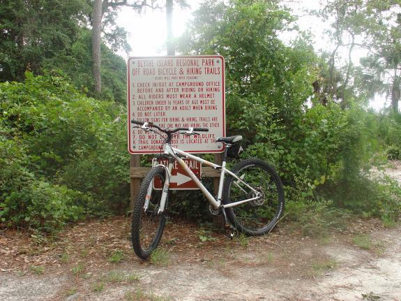 Gary Fisher X-Caliber: A mountain bike leaning against a sign for Blowing Island Regional Park, which provides information about off-road bicycle and hiking trails. The sign features rules and guidelines for park visitors, surrounded by lush greenery and a natural setting.
