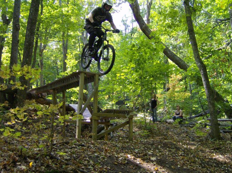 Giant AC: A mountain biker jumping off a wooden ramp in a forested area, surrounded by greenery and fallen leaves. Two spectators watch from the side as the biker is mid-air, showcasing an action-packed moment in nature.
