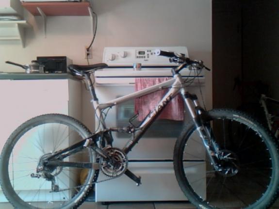 Giant Anthem 1: A mountain bike leaning against a kitchen counter with an oven in the background. The bike has a silver and black frame, thick tires, and visible gears. There are kitchen items on the counter and a red dish towel hanging from the oven.