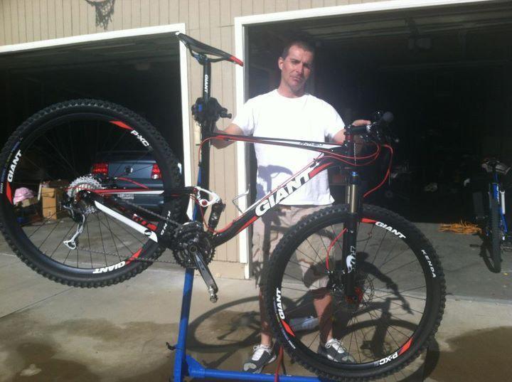 Giant Anthem 2: A person holding a mountain bike in a garage setting. The bike is elevated on a stand and features a prominent "Giant" logo. The individual is wearing a white shirt and shorts, standing in front of two open garage doors with various items visible in the background. The scene is well-lit, suggesting a sunny day.