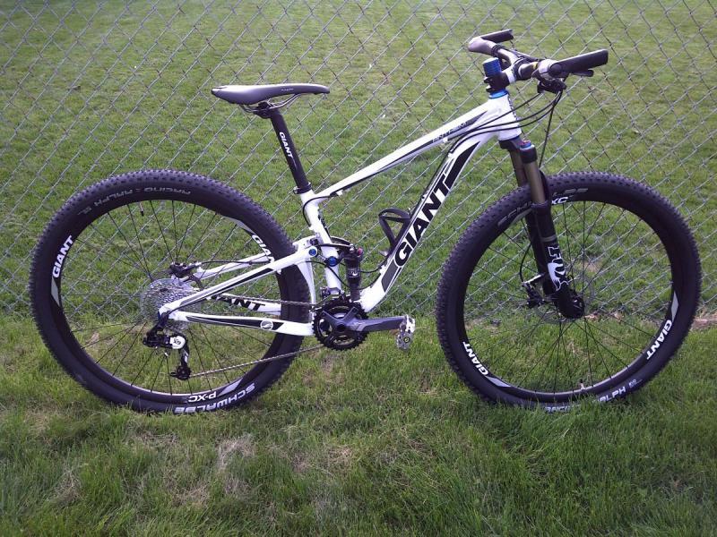 Giant Anthem X 29er 1: A white and black mountain bike leaning against a chain-link fence, set against a green grassy background. The bike features wide tires, a sturdy frame, and a front suspension fork, with visible brand logos on the frame and wheels.