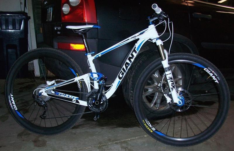Giant Anthem X 29er 3: A white mountain bike with blue accents and the brand name "Giant" prominently displayed on the frame, positioned in a garage next to a vehicle. The bike features thick tires, a suspension system, and a narrow seat, suggesting it is designed for off-road cycling.