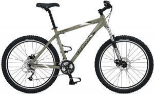 A mountain bike in a khaki color featuring knobby tires, a sturdy frame, and both front and rear disc brakes. The bike is equipped with a multi-speed gear system and a comfortable saddle, designed for off-road riding.