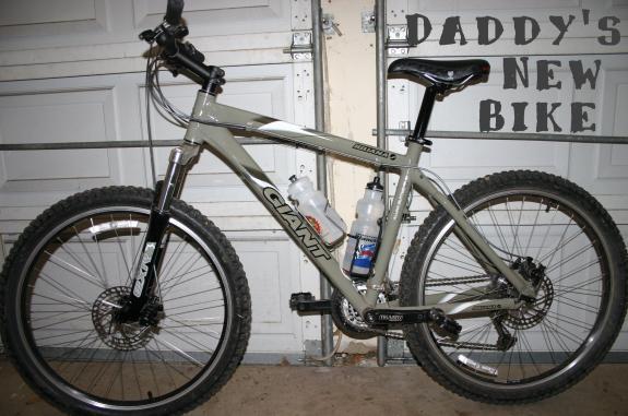Giant Iguana: A mountain bike leaning against a garage wall with two water bottles mounted in the frame. The bike is a gray Giant model with thick tires, a front suspension fork, and a black seat. In the background, there is text on the wall that reads "DADDY'S NEW BIKE."