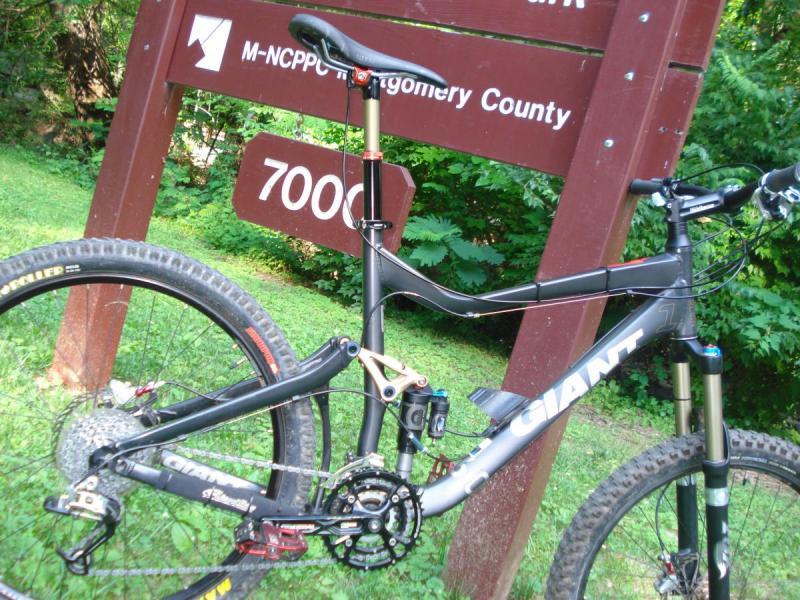 Giant Reign 1: A mountain bike is leaning against a wooden sign that reads "M-NCPPC Montgomery County" with the number "7000" visible. The background features lush greenery, indicating an outdoor environment suitable for biking. The bike has distinct features, including thick tires and a sturdy frame, suggesting it is designed for rugged terrain.