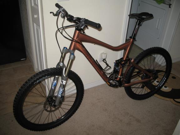 Giant Reign 1: A mountain bike with a bronze frame and large, knobby tires is positioned beside a wall in a room with light-colored flooring. The bike features front suspension forks and a black handlebar with grips, indicating it is designed for off-road use. In the background, a doorway and a laundry area are visible.