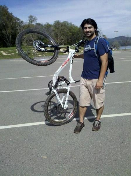 Giant Reign 2: A man stands on a paved road, holding a bicycle with one wheel lifted off the ground. He has long black hair and is wearing a blue t-shirt, beige shorts, and brown shoes. A backpack is slung over one shoulder. In the background, there are trees and a distant view of hills under a clear blue sky.