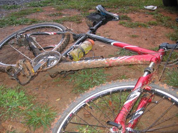 Giant Rincon: A red and black mountain bike lying on muddy ground, partially covered in dirt, with a water bottle secured in the frame. The scene depicts wet grass and a gravel area in the background, indicating a possibly muddy or challenging biking environment.