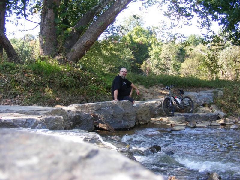 Giant Rincon: A person sitting on a rock by a stream, with a bicycle parked nearby. The scene is surrounded by lush greenery and trees under a clear sky, creating a peaceful natural setting.