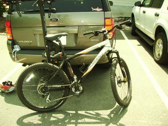 Giant Rincon: A mountain bike parked beside an SUV in a parking lot, with the bike's front wheel slightly turned and a bike rack mounted on the back of the vehicle. The setting is sunny, and the surrounding vehicles are parked in adjacent spaces.