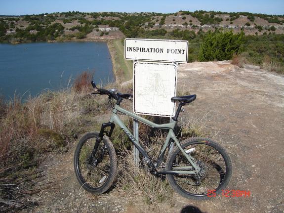 Giant Rincon: A mountain bike parked next to a sign that reads "Inspiration Point," overlooking a tranquil body of water surrounded by greenery and hills. The scene captures a peaceful outdoor setting, ideal for enjoying nature and cycling.