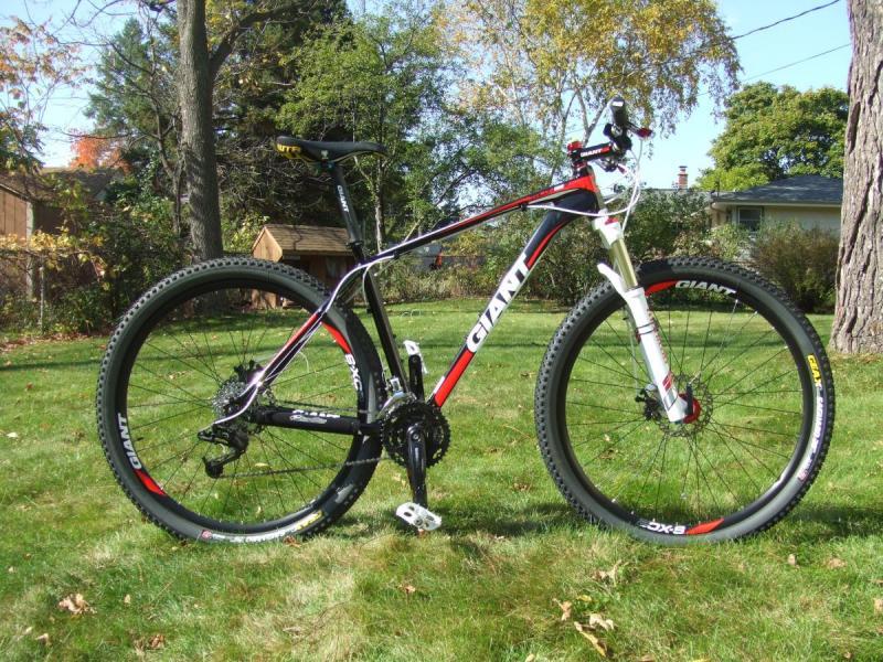 Giant Talon 29er: A black and red mountain bike parked on green grass, featuring a lightweight frame, front suspension, and thick, knobby tires. The bike has branding from the manufacturer "Giant" visible on its frame and wheels, with a scenic background including trees and a small shed.