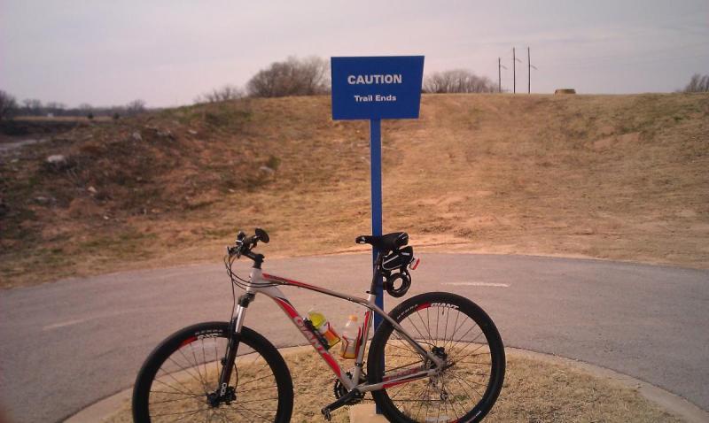 Giant Talon 29er: A mountain bike parked on a pathway near a blue sign that reads "CAUTION Trail Ends," with a grassy area and power lines visible in the background. The scene is overcast, indicating a cloudy day.
