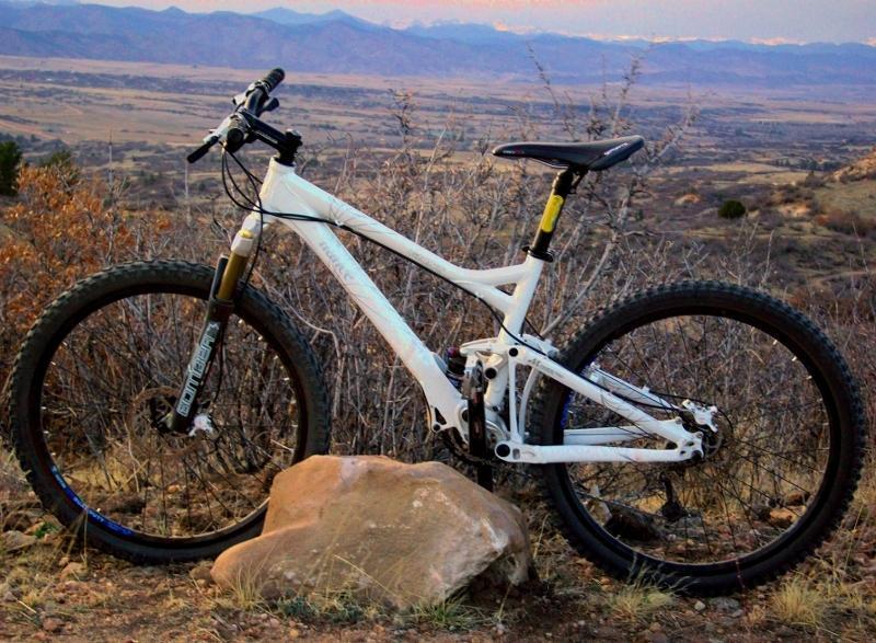 Giant Trance 1: A white mountain bike resting on a rock, with a scenic view of rolling hills and distant mountains in the background. The landscape features dry brush and an expansive valley, under a soft evening light.