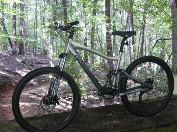 Giant Trance 2: A silver mountain bike parked on a gravel path, surrounded by a dense forest with tall trees and green foliage.