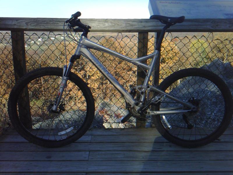 Giant Trance: A silver mountain bike leaning against a wooden railing with a chain link fence in the background, set against a backdrop of fall foliage. The bike features thick tires and a dual suspension system, suggesting it's designed for off-road riding.