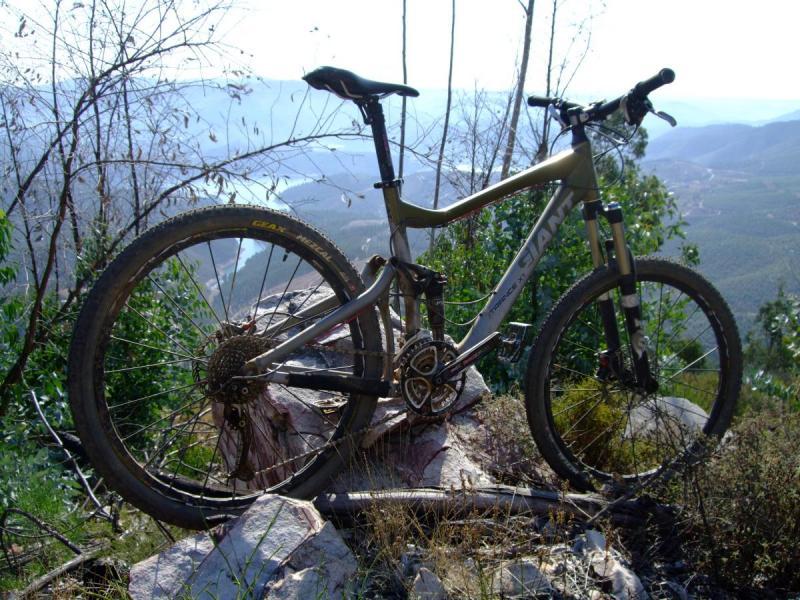 Giant Trance X1: A mountain bike resting on a rocky outcrop, surrounded by sparse vegetation and overlooking a scenic landscape of rolling hills and distant mountains under a clear blue sky.
