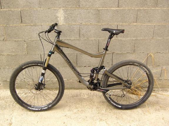 Giant Trance X1: A mountain bike with a metallic gray frame, front suspension, thick tires, and a black seat, positioned against a concrete block wall.