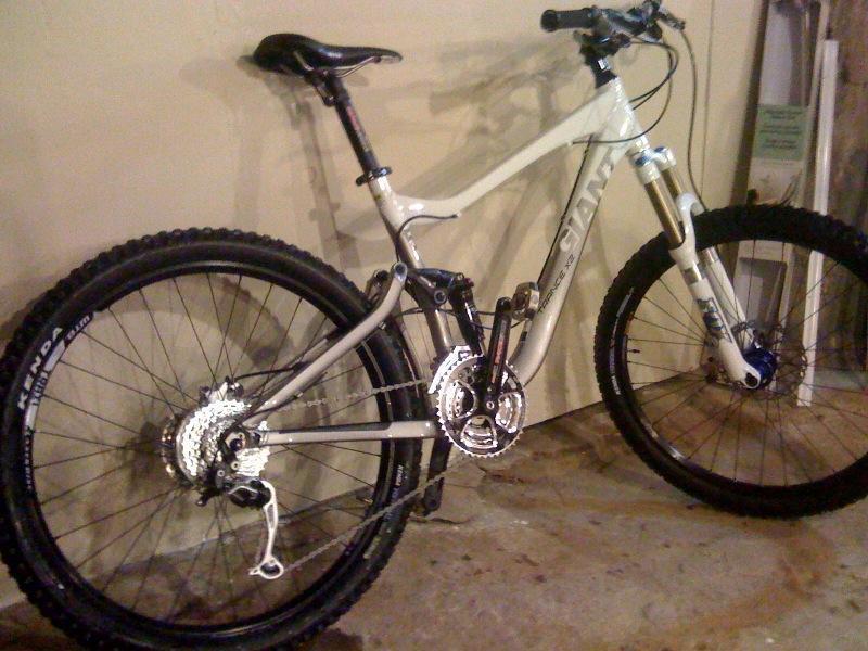 Giant Trance X2: Alt text: A white mountain bike with thick black tires is leaning against a beige wall. The bike features a rear suspension system, multiple gears, and a sturdy frame designed for off-road riding. The background shows a simple indoor setting.