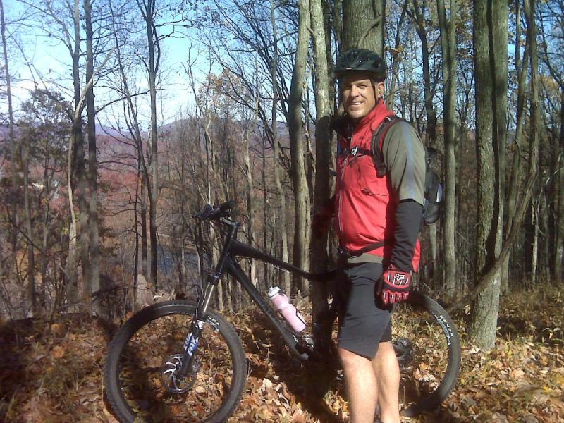 Giant Trance X2: A mountain biker stands next to his bike on a wooded trail during autumn. He wears a black helmet and red vest, with a water bottle attached to the bike. The background features trees with fall foliage and a clear blue sky.