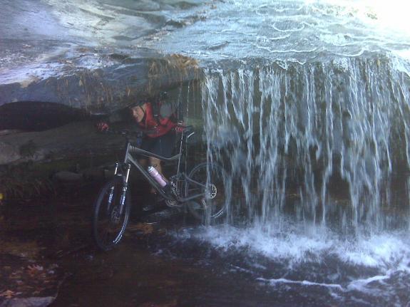 Giant Trance X2: A mountain biker positioned just beneath a waterfall, partially obscured by the cascading water. The cyclist wears a red jacket and is crouched on a bike as water flows over a rocky overhang above. The scene captures the dynamic interplay between adventure and nature in a lush, green environment.
