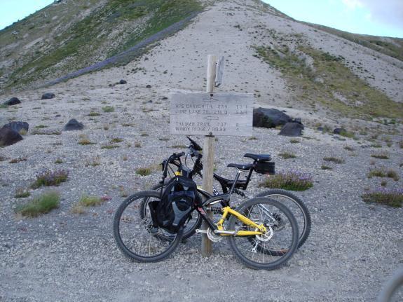 A pair of mountain bikes lean against a wooden trail sign indicating various hiking distances and destinations, set against a backdrop of a rocky hillside with patches of greenery.