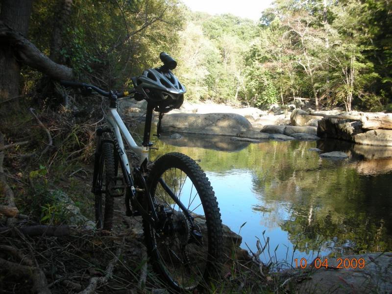 Giant Yukon FX: A mountain bike leaning against a tree near a calm river surrounded by lush greenery and rocky banks. The photo captures a peaceful outdoor scene, ideal for cycling enthusiasts.