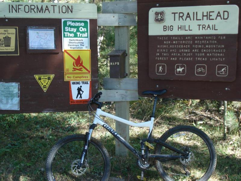 Giant Yukon FX: An information sign at the trailhead for the Big Hill Trail, featuring multiple guidelines including no campfires and staying on the trail, with a mountain bike leaning against it. The sign displays various symbols related to trail usage and recreational activities.