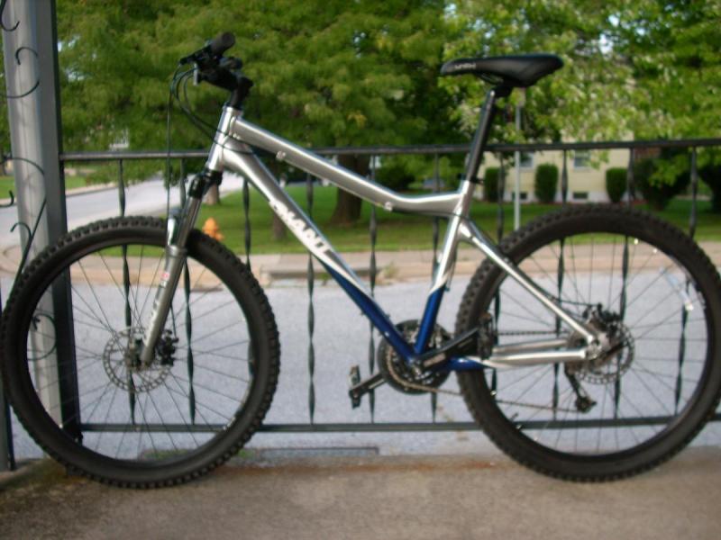 Giant Yukon: A silver and blue mountain bike is leaning against a metal railing. The bike has thick, treaded tires and a simple design with a focus on durability. In the background, a residential street and lush green trees are visible, indicating an outdoor setting.