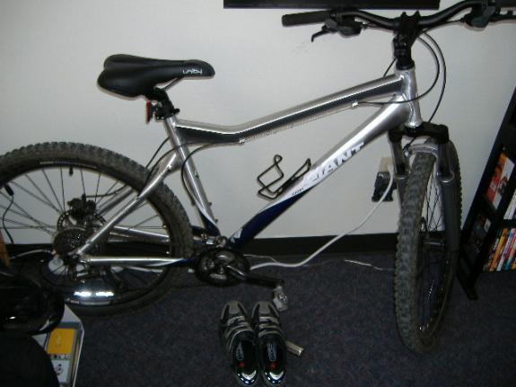 Giant Yukon: A silver mountain bike leaning against a wall, with a pair of black and gray cycling shoes placed beside it on a carpeted floor. The bike features chunky tires and a visible gear system, with a water bottle holder attached to the frame. In the background, there is a bookshelf filled with DVDs.