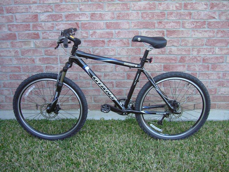 Giant Yukon: A black mountain bike with a sleek frame and thick tires, parked on green grass beside a textured red brick wall. The bike features a single seat, handlebars with brake levers, and a visible gear system.