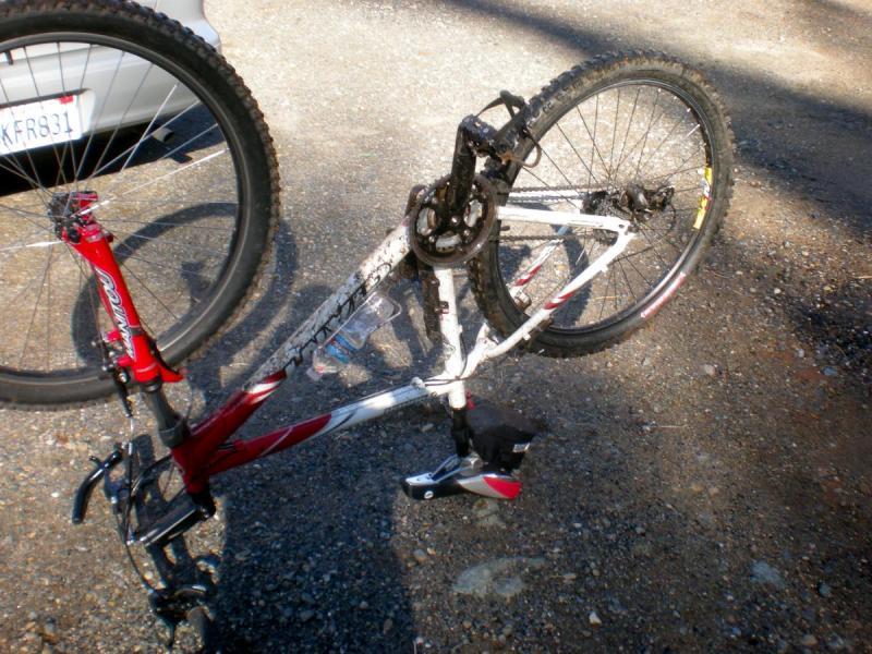 Giant Yukon: A bicycle lying on its side with the rear wheel elevated, showing the bike frame, chain, and pedals. The bike features a red and white color scheme and is positioned on a gravel surface. A parked car is visible in the background.