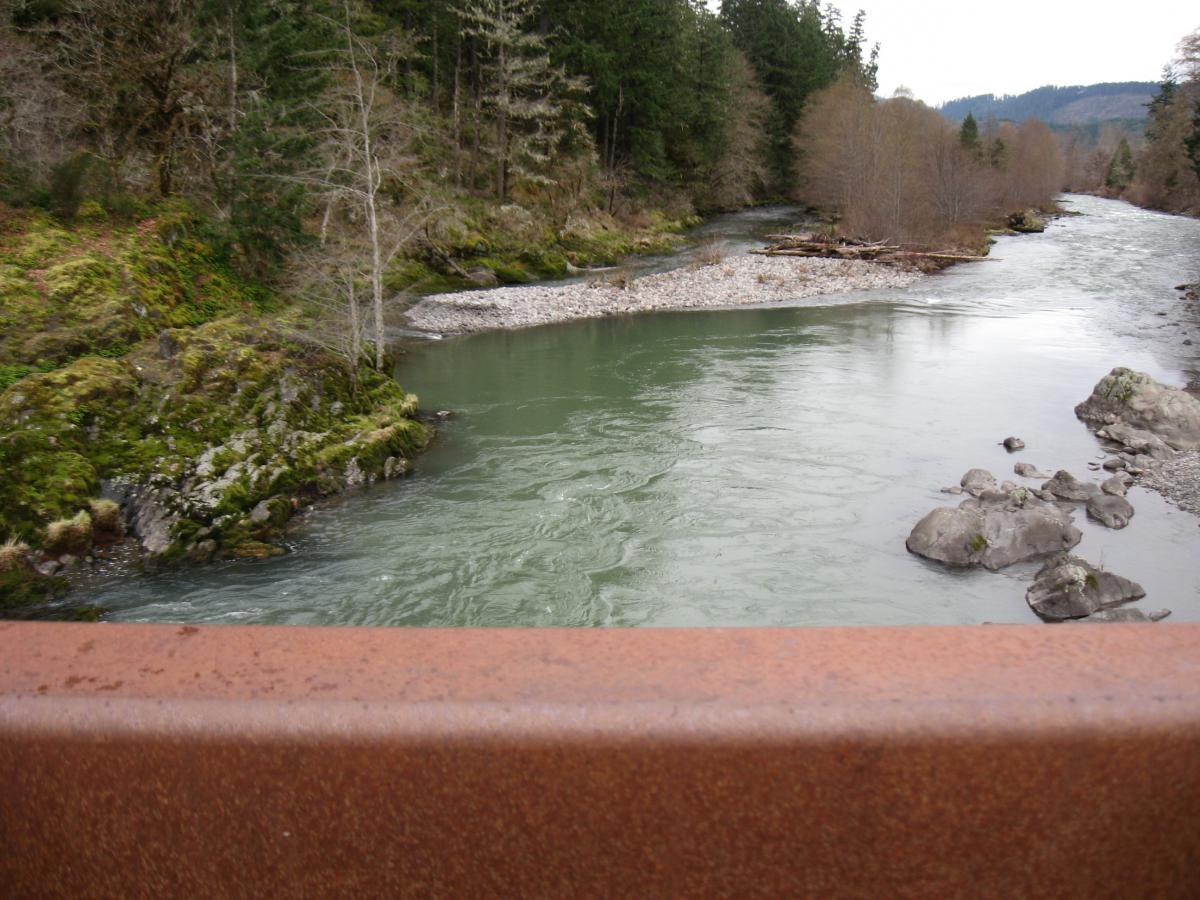 A scenic view of a river winding through rocky banks and lush greenery, captured from above. The water appears calm and flows smoothly, surrounded by trees in the background, hinting at a tranquil, natural setting. Larison Rock mountain bike trail.