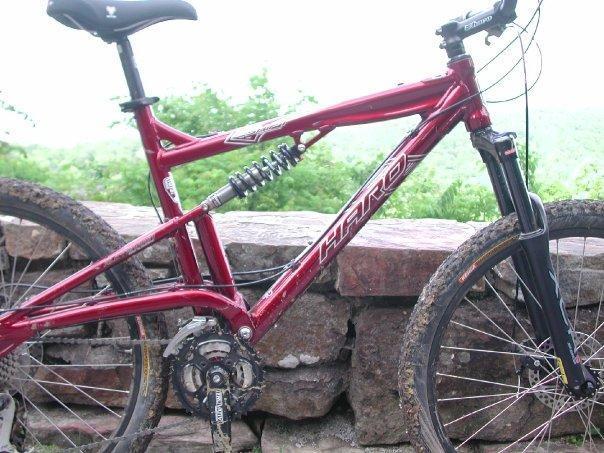 Haro Shift R1: A close-up view of a red mountain bike with suspension, resting against a stone wall. The bike features thick tires and a gear system, surrounded by a natural green landscape in the background.