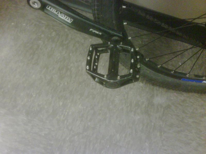 Haro Shift R5: Close-up of a black bicycle pedal attached to the crank arm, with a textured surface for grip, set against a dark, patterned floor. The pedal is part of a bicycle, partially visible in the background.
