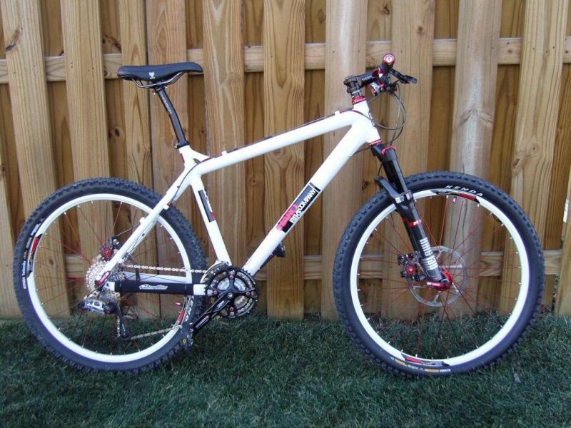 Haro V4: A white mountain bike with red and black accents, featuring a sturdy frame, wide tires, and front suspension. The bike is positioned upright on grassy ground with a wooden fence in the background.