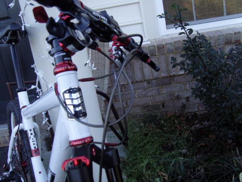Haro V4: A close-up view of a white mountain bike's handlebars and front fork, featuring red accents and cables, positioned near a brick wall and some greenery. In the background, decorative lights are visible on the porch.