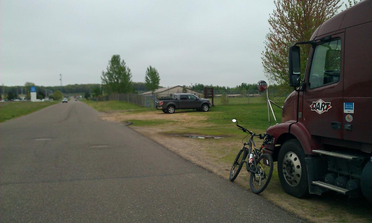 A large maroon truck is parked along a gravel road. Beside it, a bicycle is leaning against the truck. The scene includes trees on the right along a fenced area, with a gray pickup truck parked further down on the left. The sky is overcast, suggesting a cloudy day. Lowes Creek mountain bike trail.