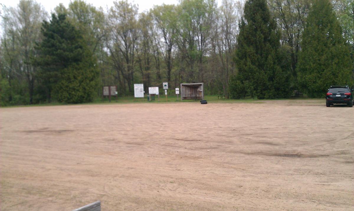 A sandy, open area surrounded by trees, with signage in the background providing information. A small shelter and a car parked on the right side of the image are visible. The scene is overcast, suggesting a calm, quiet environment. Lowes Creek mountain bike trail.