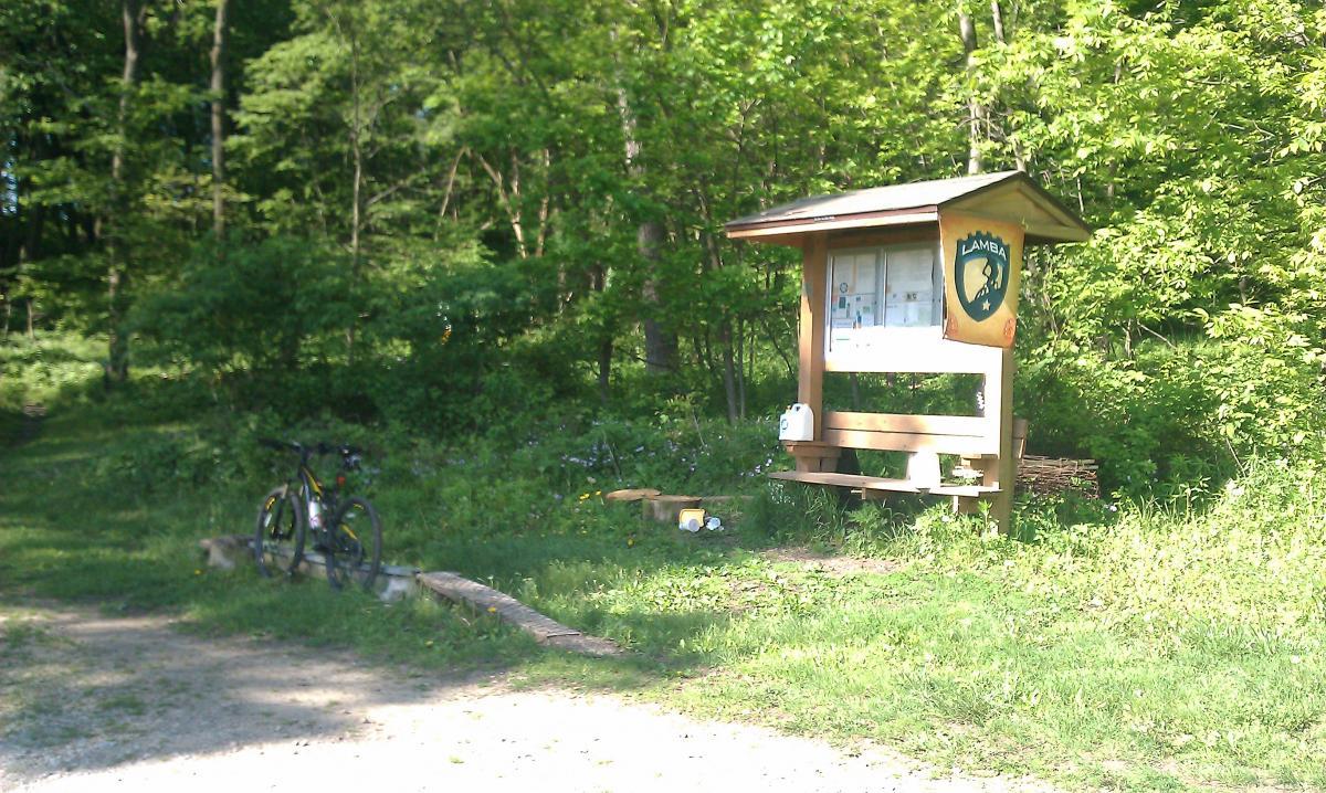 A wooden information kiosk at the edge of a forested area, surrounded by green foliage. A bicycle is parked nearby, and a small bench is visible in front of the kiosk, which displays maps and trail information. The scene is bathed in natural sunlight, indicating a pleasant day for outdoor activities. Beverly Park mountain bike trail.