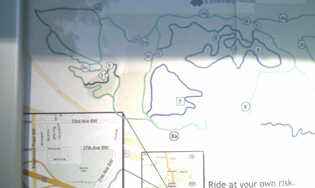 A detailed map indicating various trails, routes, and difficulty levels within a park area. The map features blue and green lines representing different trail paths, along with numbered markers for reference. A smaller inset map shows surrounding streets and landmarks, with a cautionary note at the bottom advising users to ride at their own risk. Beverly Park mountain bike trail.