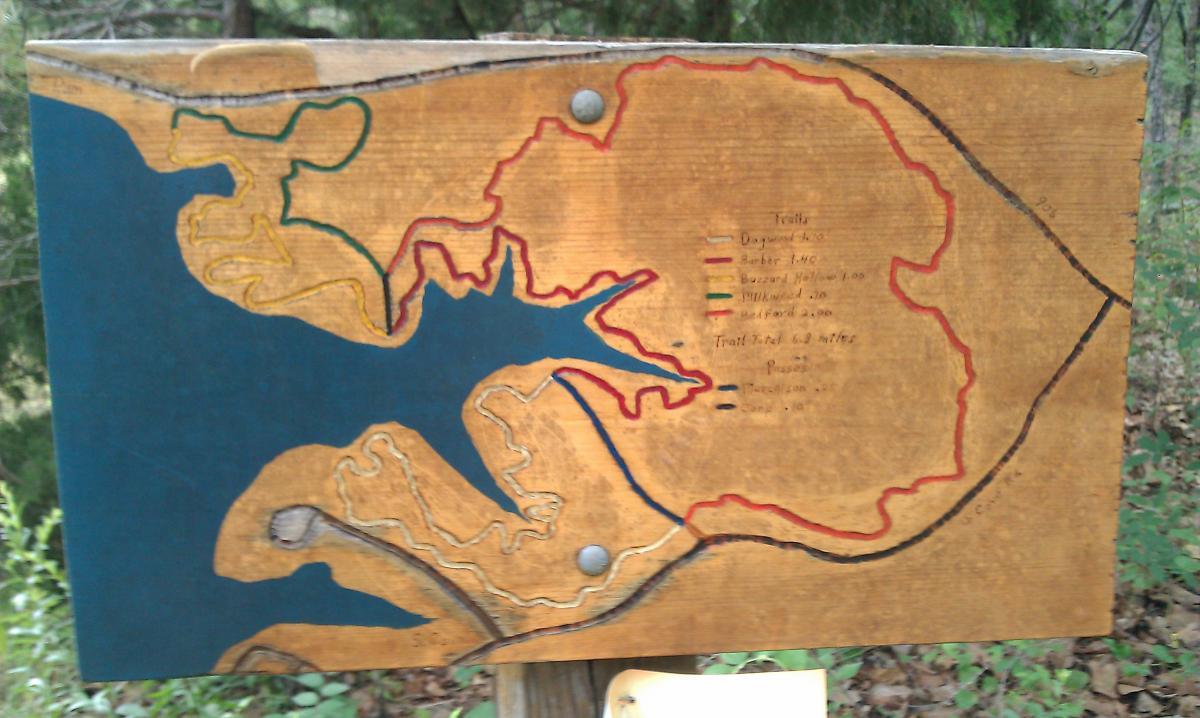 A wooden trail map sign displaying various hiking trails around a body of water. The map is illustrated with colored lines indicating different trails: yellow for Dagger's Loop, red for Barber Trail, and green for Buzzard Hollow, along with distances for each. The background features a blue area representing the water, and additional information about trail totals and access points is provided on the sign. The sign is surrounded by trees and natural foliage. Barber Hills Trail at Pat Mayse Lake mountain bike trail.