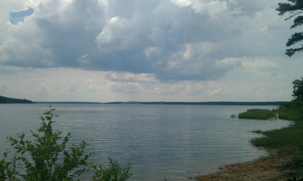 A serene lake view under a partly cloudy sky, with gentle ripples on the water's surface. Lush greenery frames the foreground, while distant hills can be seen along the horizon. Barber Hills Trail at Pat Mayse Lake mountain bike trail.