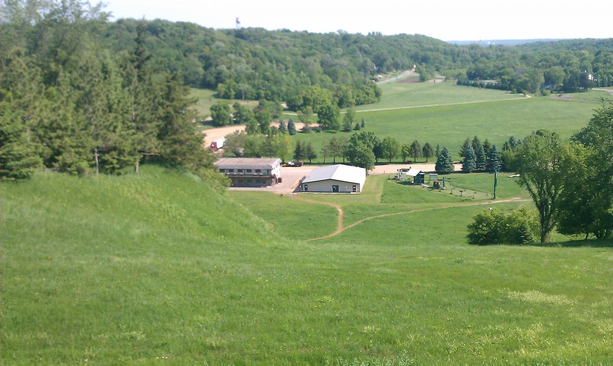 A scenic view of a green landscape featuring rolling hills, a cluster of buildings, and a winding dirt path. The foreground is dominated by lush grass, while the background showcases trees and hills under a clear sky. The buildings include a larger white structure and a multi-story building, surrounded by fields and greenery. Mountkato Ski Area mountain bike trail.