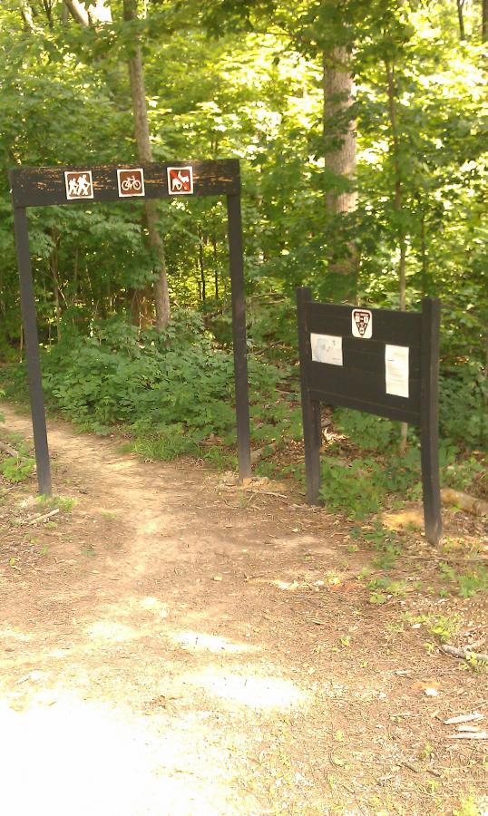 Wooded trail entrance with signs displaying icons for hiking, biking, and horseback riding. A wooden signboard with additional information is located to the right, surrounded by lush green foliage. O'bannon Woods mountain bike trail.