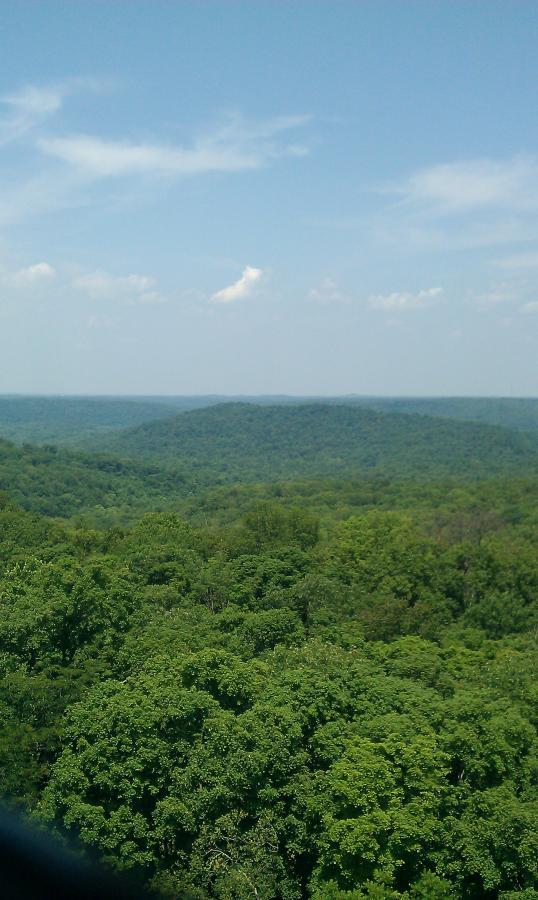 A panoramic view of lush green hills under a clear blue sky, showcasing layers of rolling landscapes and scattered clouds. O'bannon Woods mountain bike trail.